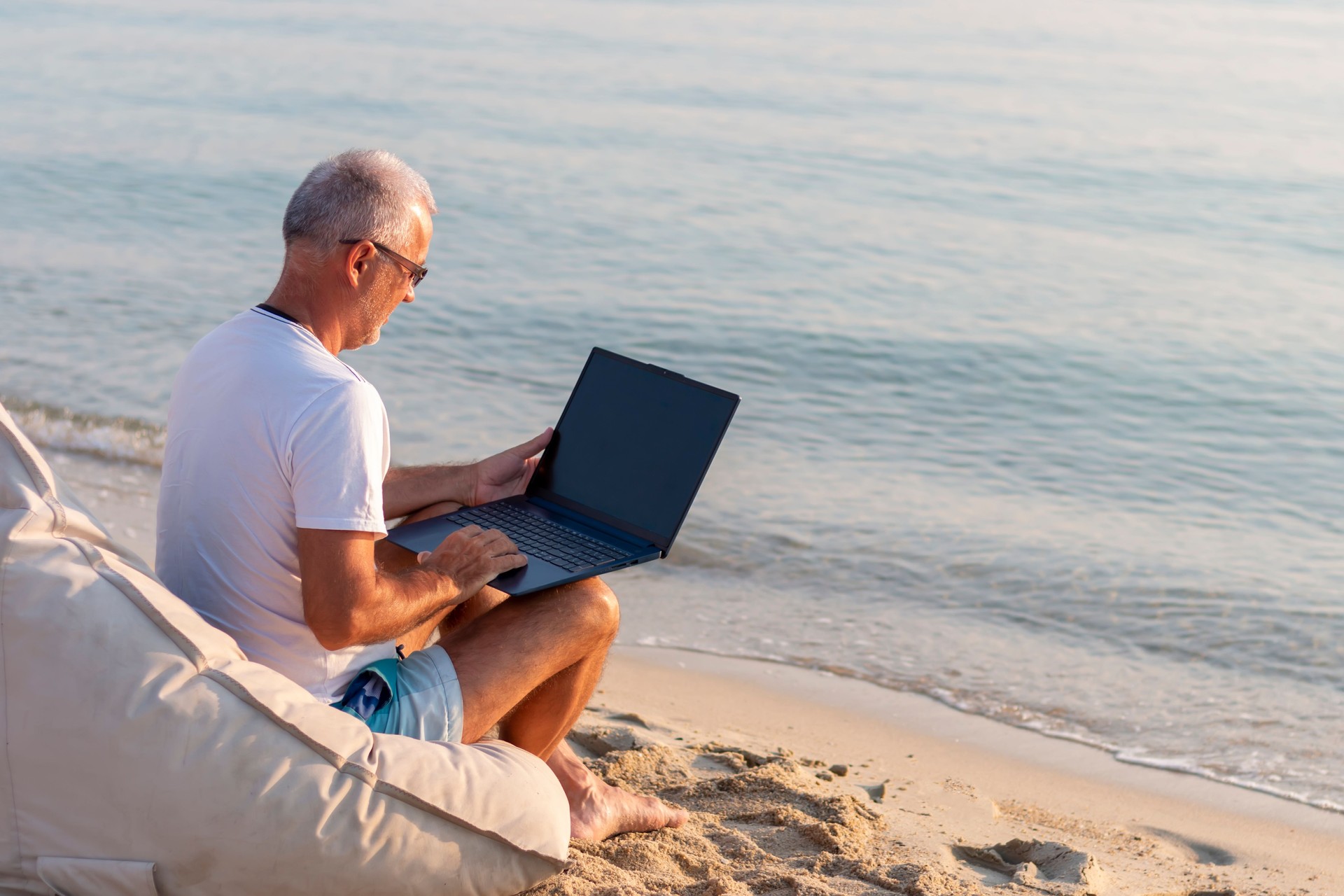 A senior male tourist in casual clothing sitting on the beach working on his laptop
